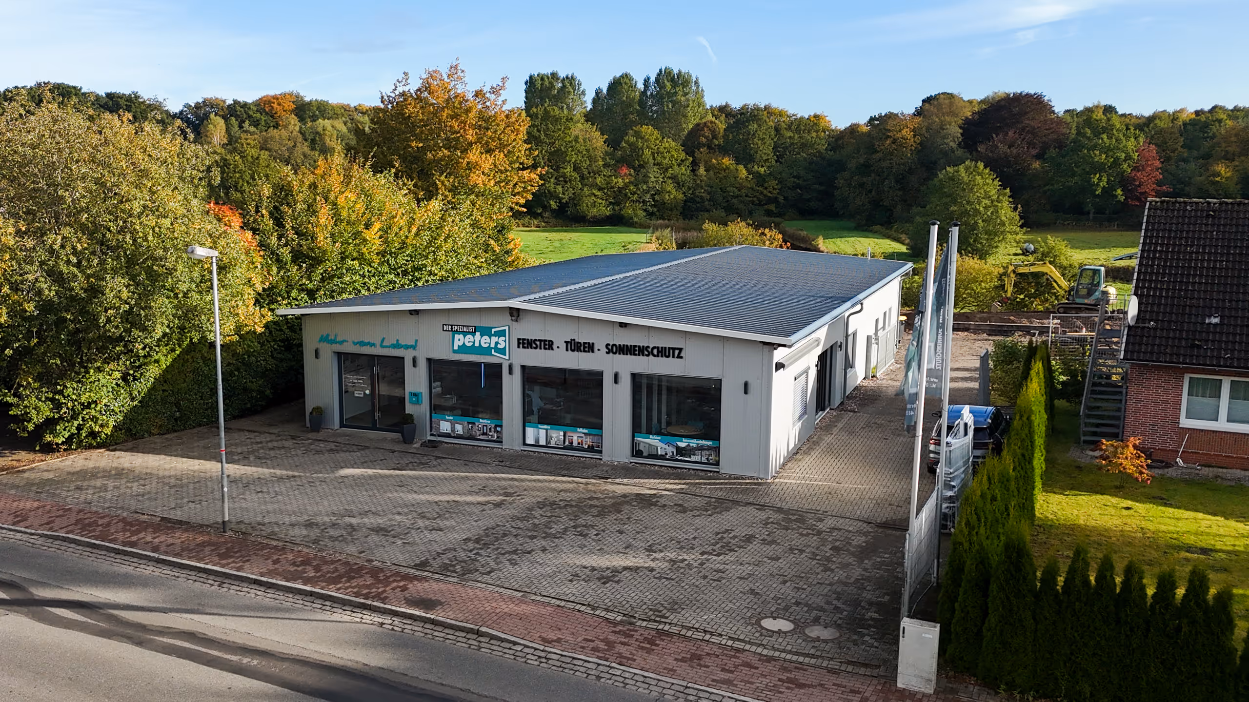 Ein flaches Gewerbegebäude mit dem Schriftzug 'peters' und Angeboten für Fenster, Türen und Sonnenschutz, umgeben von Bäumen und einer gepflasterten Fläche.
