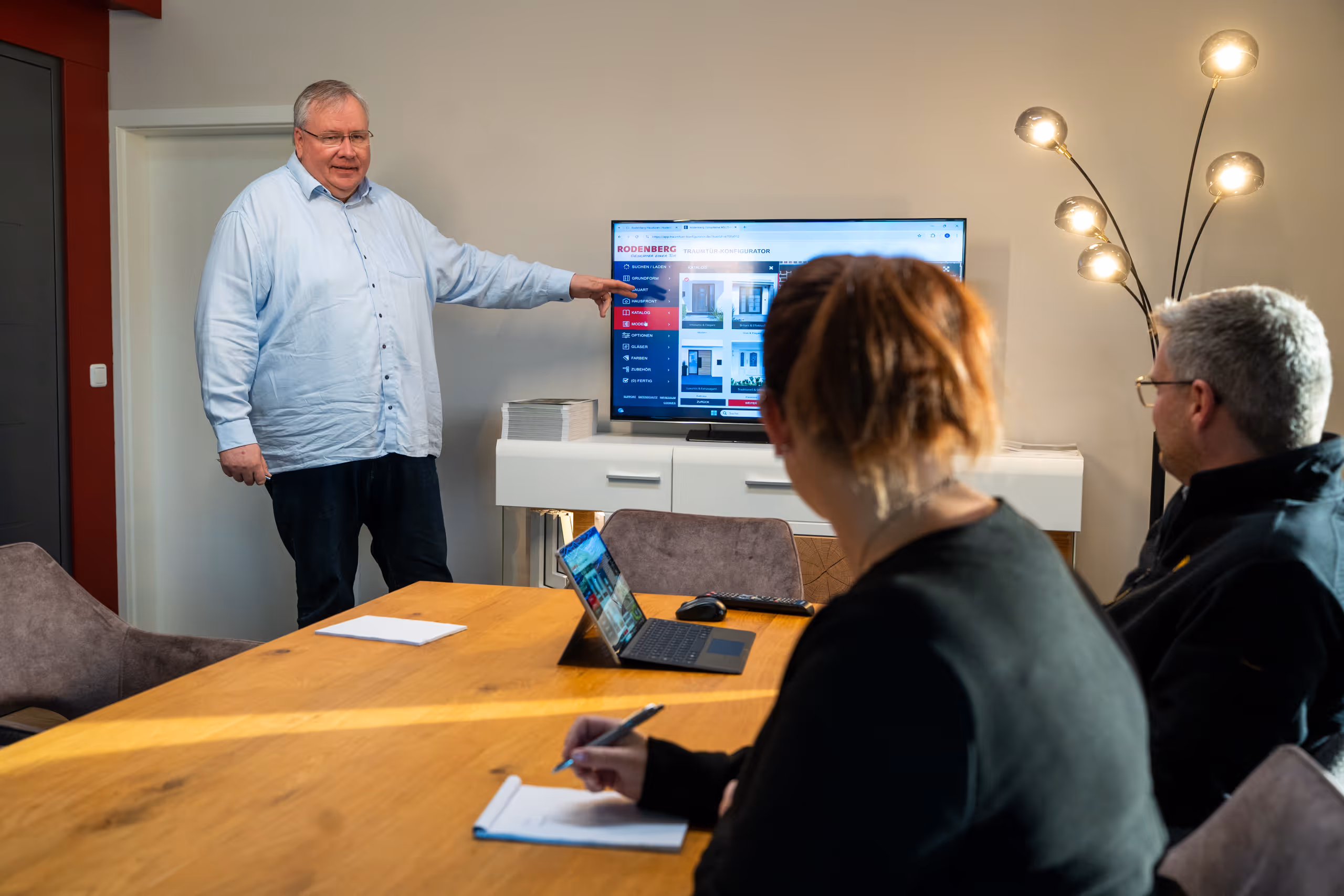 Ein Mann präsentiert eine digitale Türkonfiguration auf einem Bildschirm, während zwei Personen an einem Tisch zuhören und Notizen machen.