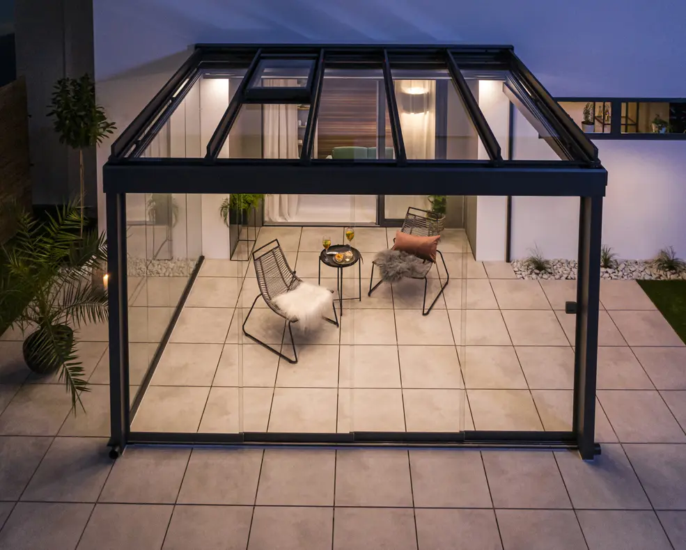 Moderner Glaspavillon mit zwei Stühlen, einem kleinen Tisch mit Getränken und Snacks, umgeben von Fliesenboden und Pflanzen.