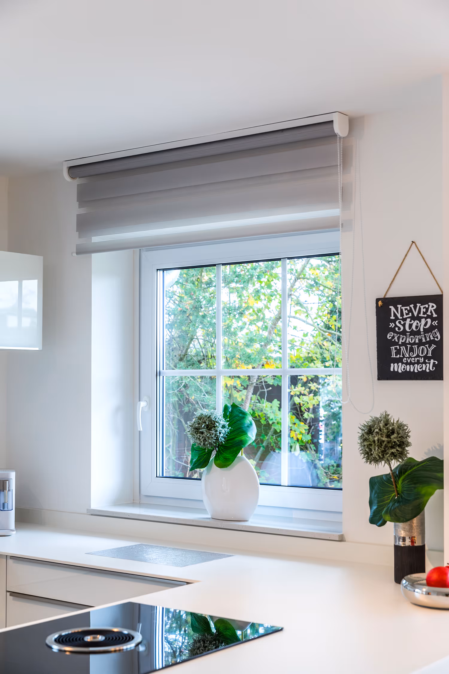 Moderne weiße Küche mit Fenster, grauer Jalousie, zwei Vasen mit grünen Pflanzen und einem Schild mit der Aufschrift ‚Never stop exploring, enjoy every moment‘.