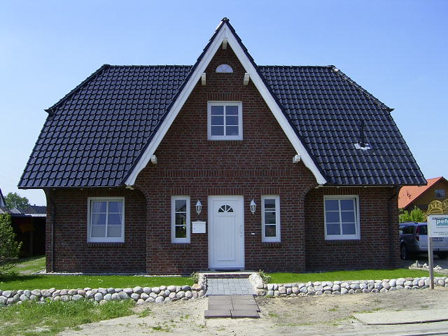 Weißen Fensterrahmen mit vier Scheiben vor einer roten Ziegelmauer, in den Scheiben spiegeln sich grüne Bäume und blauer Himmel.