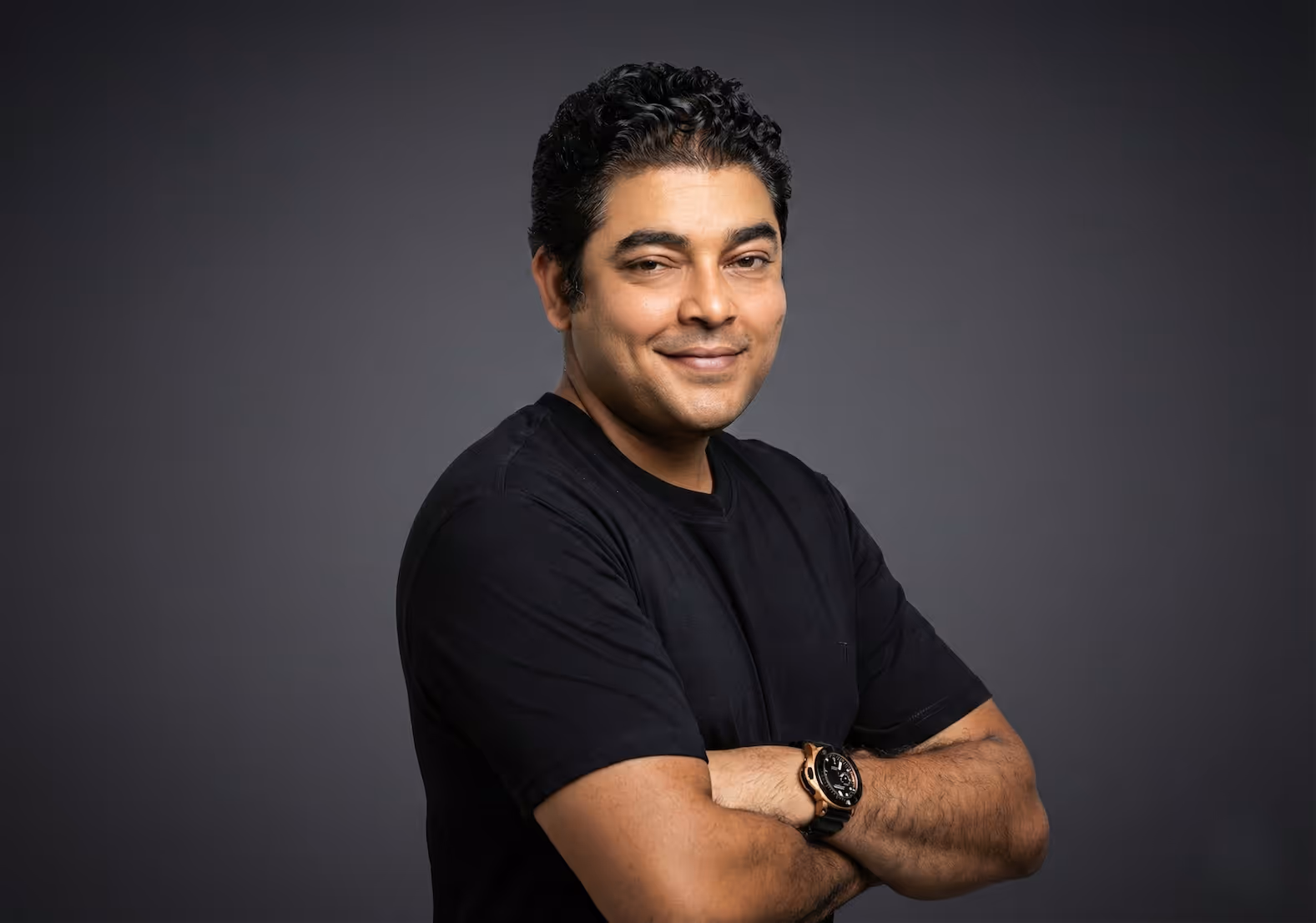Smiling man with curly hair wearing a black t-shirt and watch, standing with arms crossed against a dark gray background.