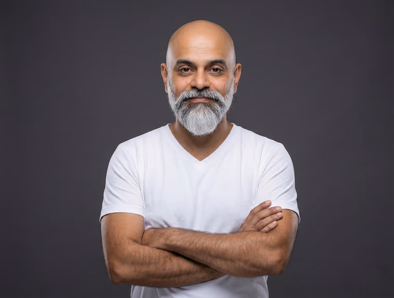 Smiling bald man with a gray beard and mustache wearing a white T-shirt, arms crossed, standing against a dark gray background.