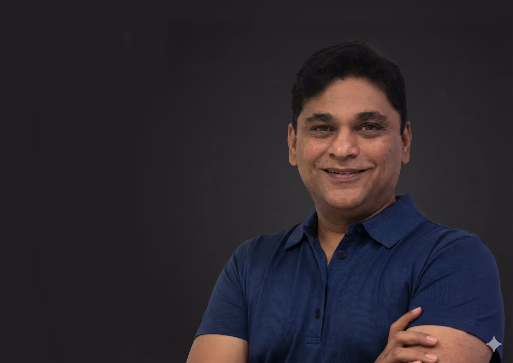 Smiling man with crossed arms wearing a blue polo shirt against a dark background.