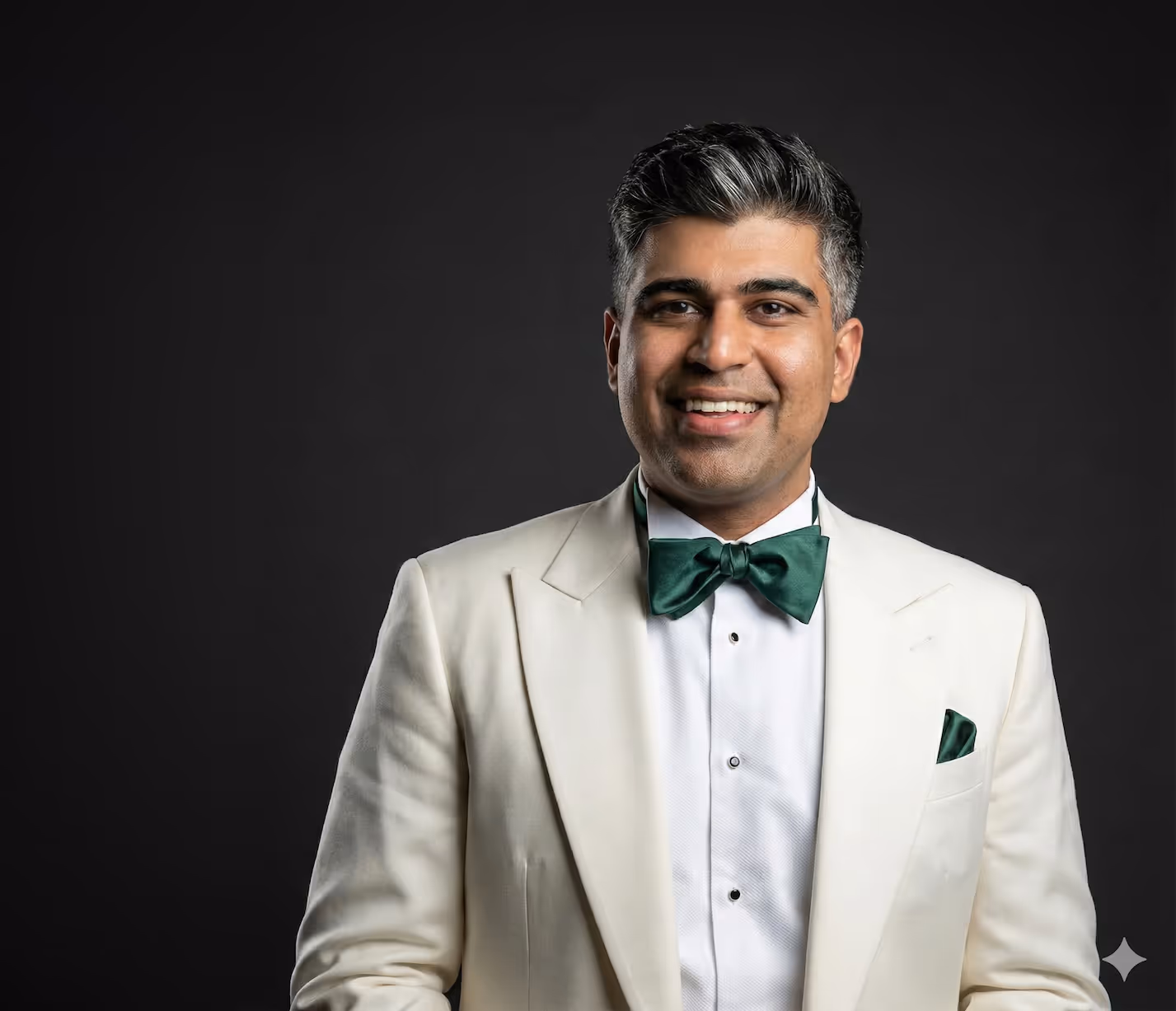 Smiling man wearing a white tuxedo jacket, white shirt, and dark green bow tie and pocket square against a dark background.