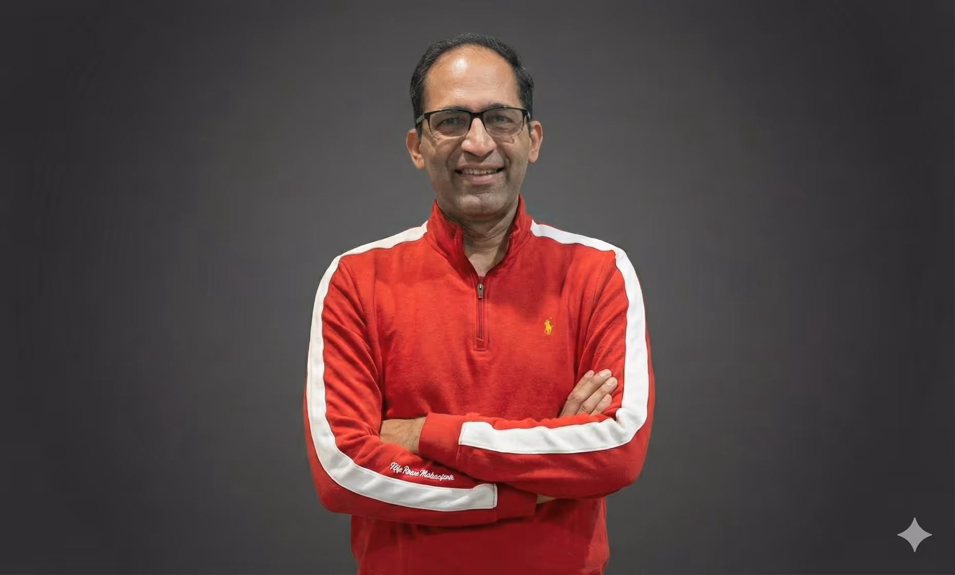 Smiling man with glasses wearing a red long-sleeve pullover with white stripes standing with arms crossed against a gray background.