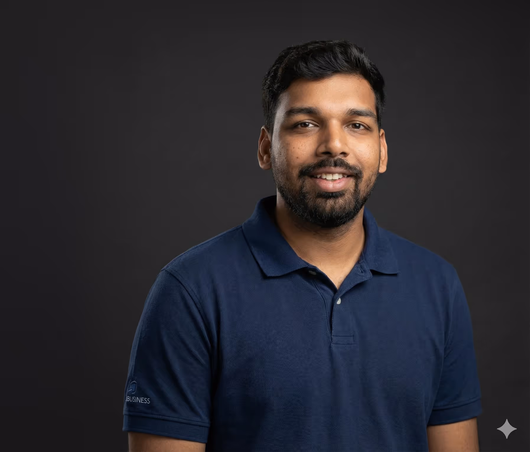 Smiling man with short black hair and a beard wearing a navy blue polo shirt against a dark background.