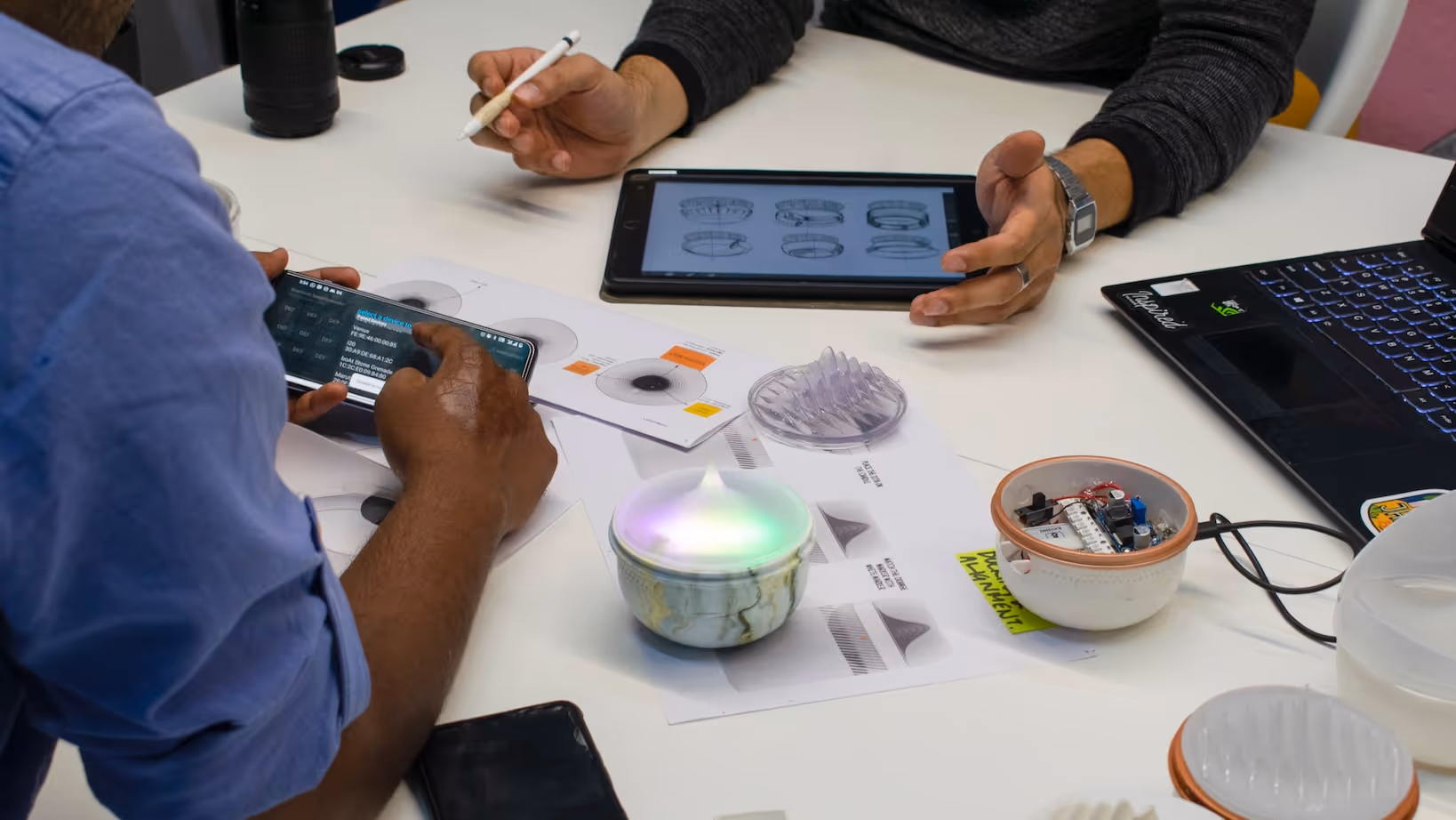 Two people working at a table with printed designs, a tablet showing sketches, a smartphone, and electronic device prototypes.