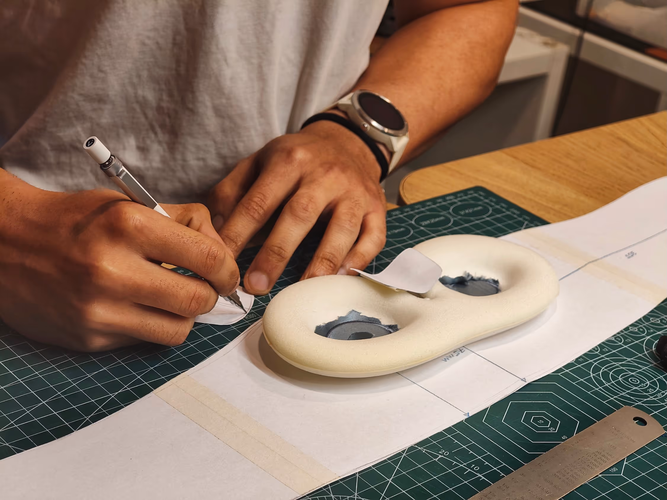 Person tracing a pattern on paper next to a foam shoe insole prototype on a green cutting mat.