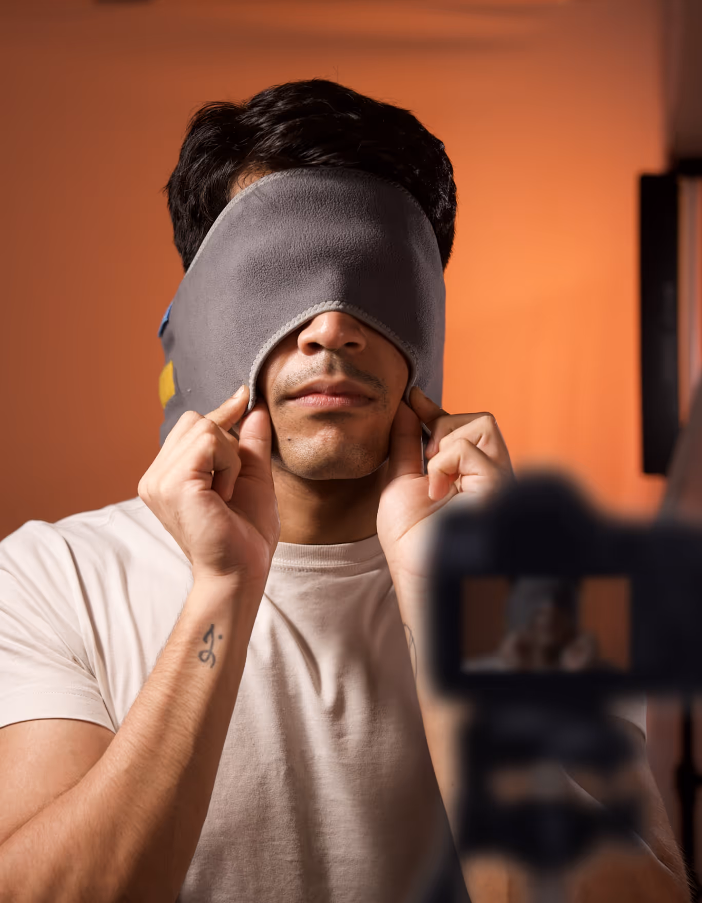 Man wearing a gray eye mask covering his eyes, adjusting it with both hands against an orange background.