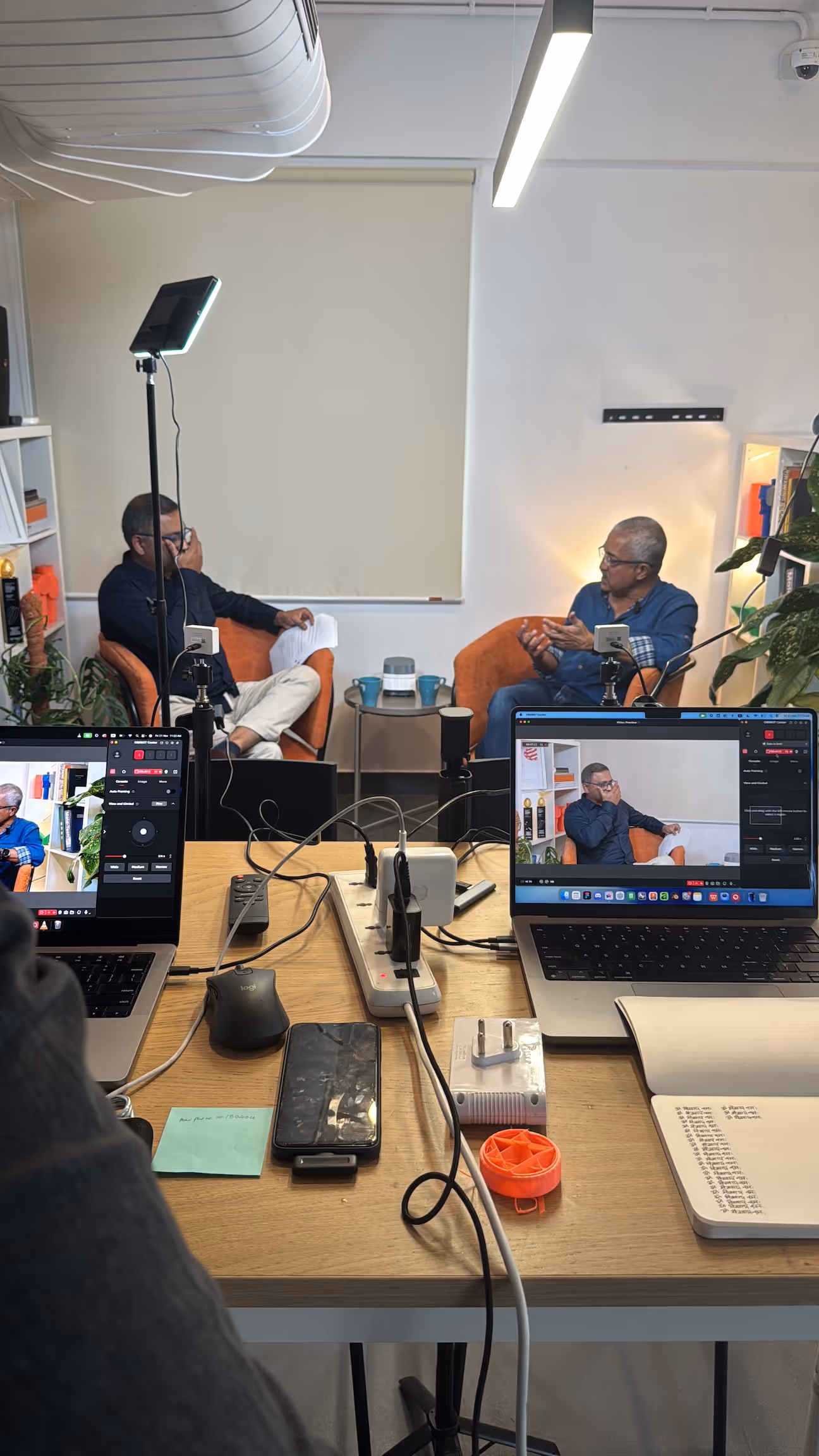 Two men seated in orange chairs having a recorded conversation with laptops and recording equipment on a desk in the foreground.