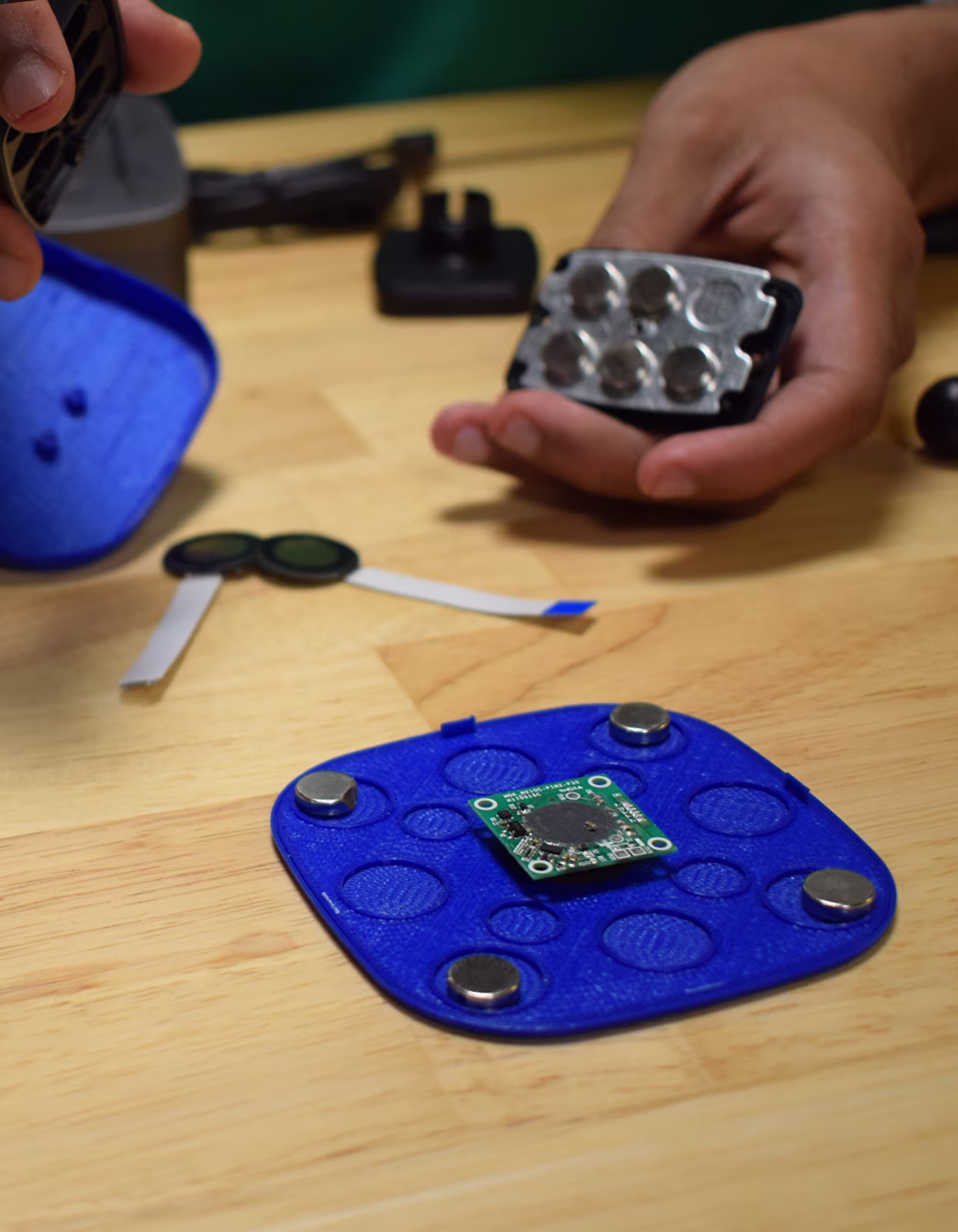 Hands assembling electronic components and 3D-printed blue casings on a wooden table.