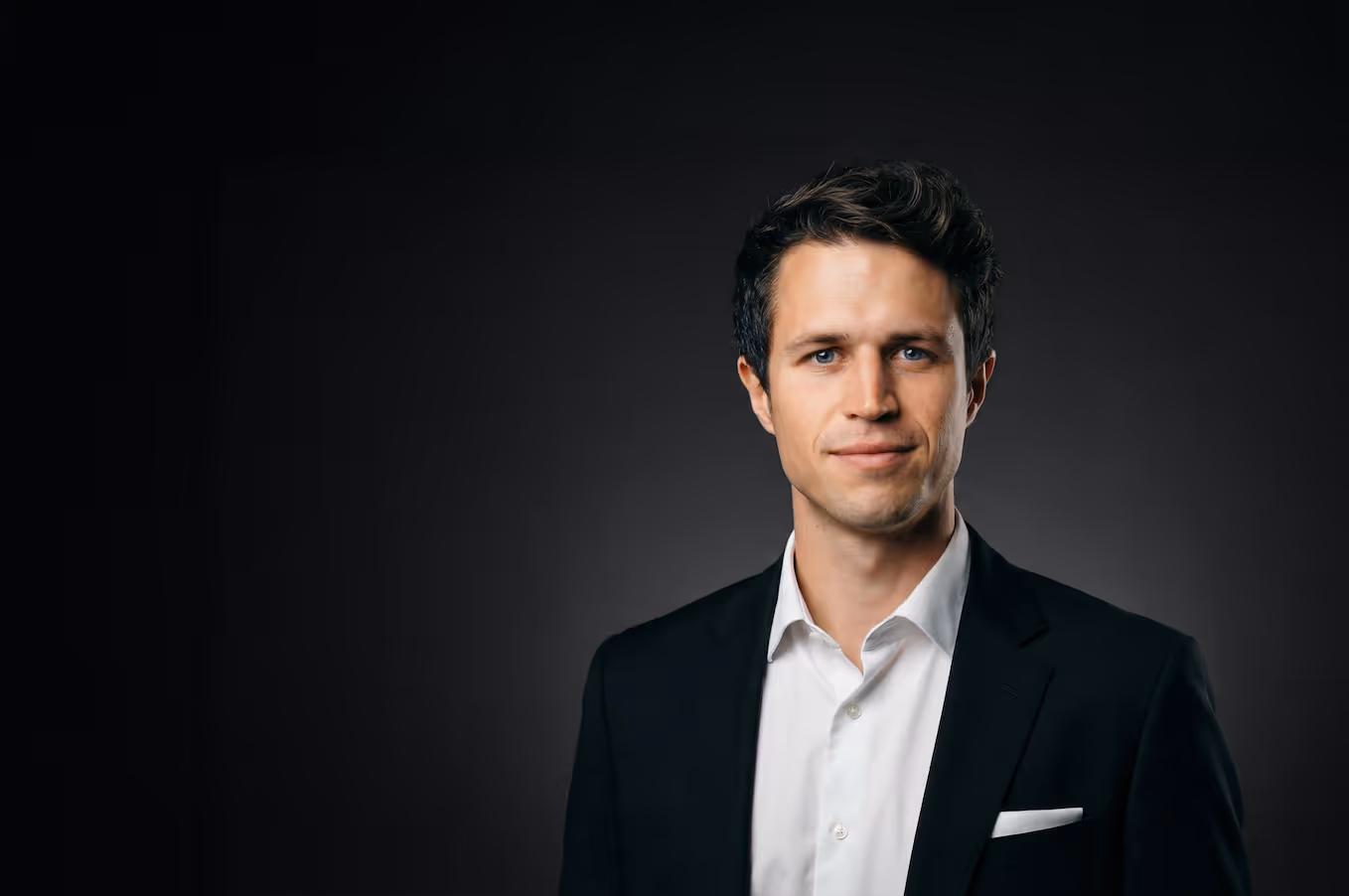 Man with short hair and beard wearing glasses on his head and a black t-shirt, smiling against a dark background.