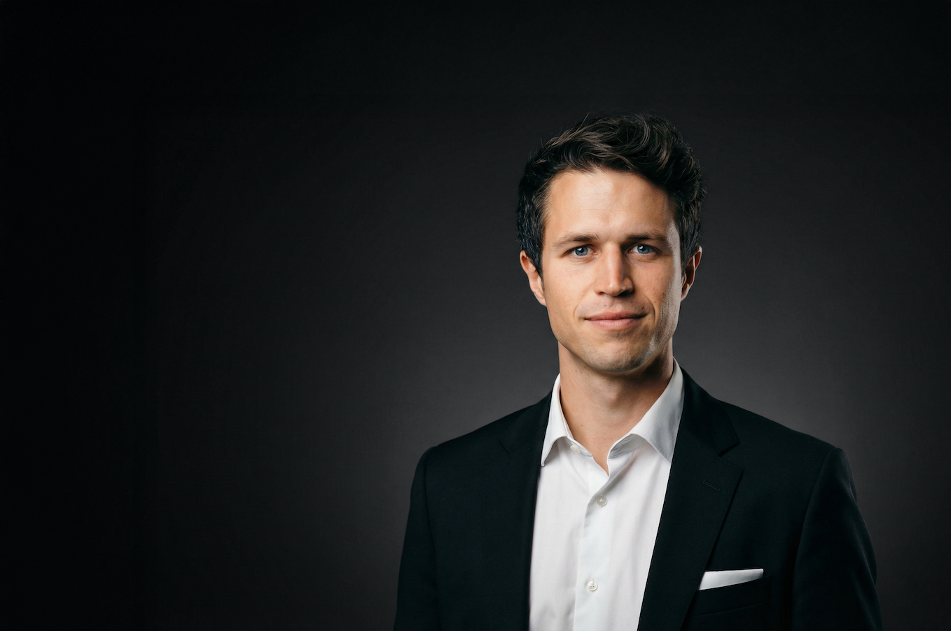 Man with short hair and beard wearing glasses on his head and a black t-shirt, smiling against a dark background.