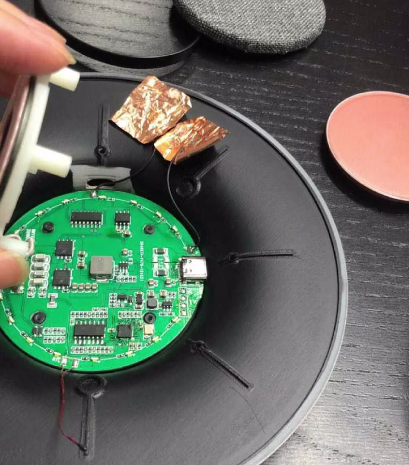 Close-up of hands assembling a black speaker with visible green circuit board and copper foil inside.