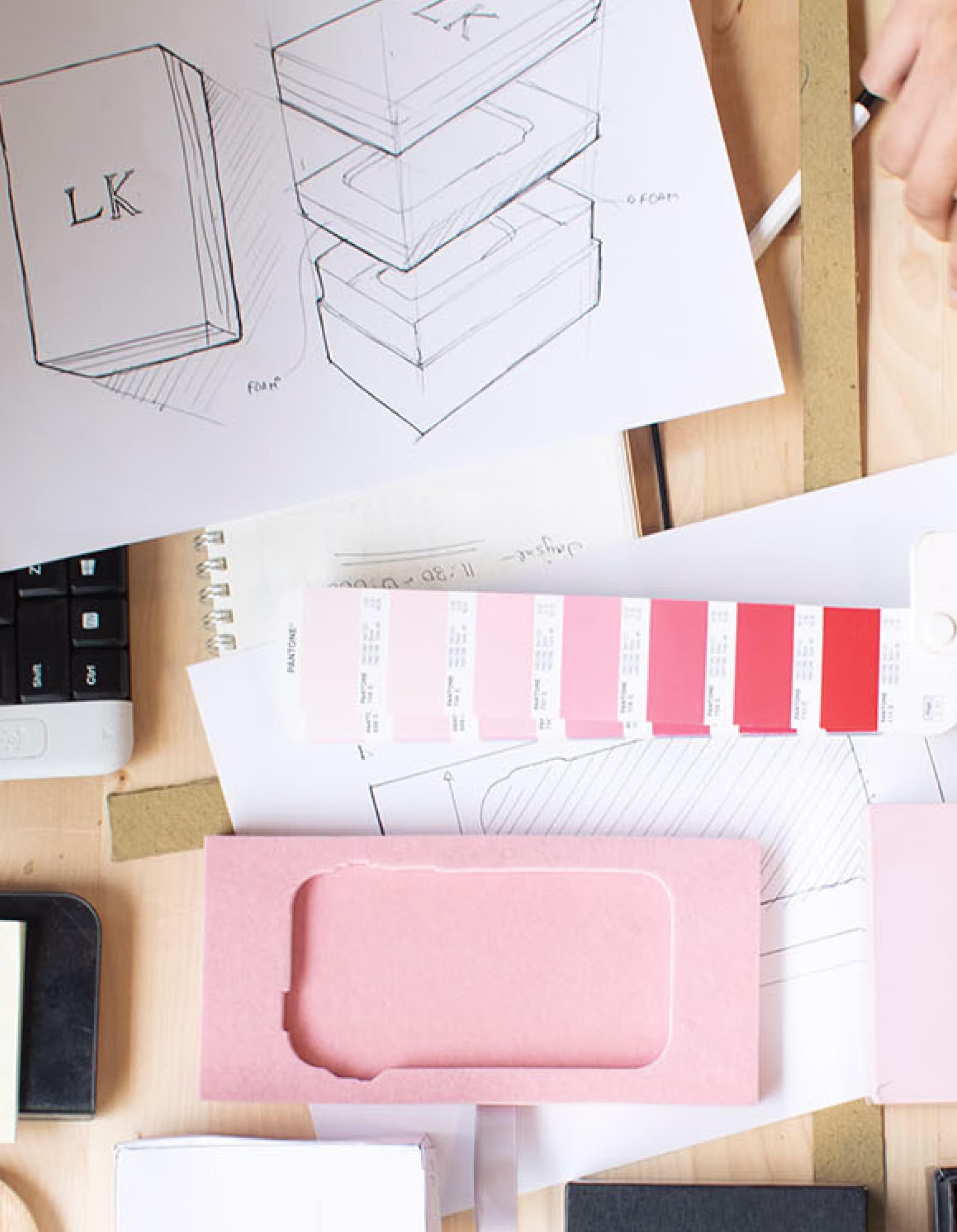 Top view of a workspace with phone packaging prototypes, color swatches, sketches, a keyboard, mouse, and a hand reaching over the table.