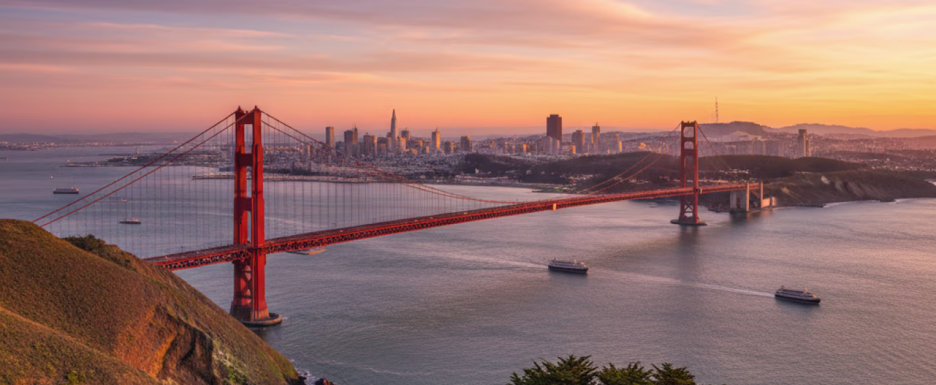 Curved coastal highway with light trails along a city skyline at sunset with a gradient orange and purple sky and birds flying.