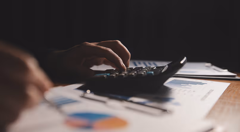 Hand pressing buttons on a calculator with financial documents and charts on a desk in dim lighting.