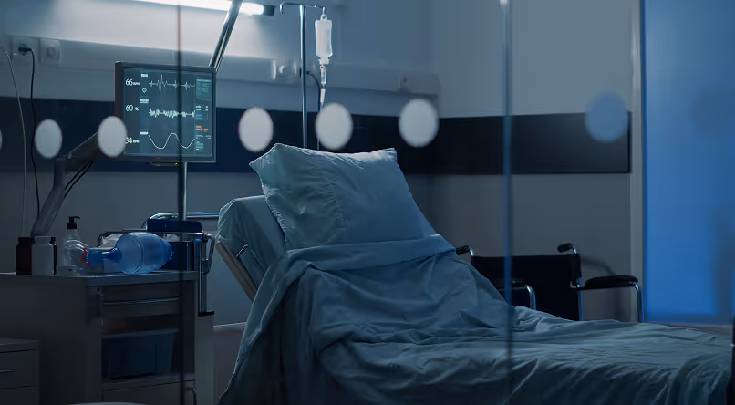Empty hospital bed with blue sheets and pillow in a dimly lit patient room, showing a medical monitor and IV drip stand.