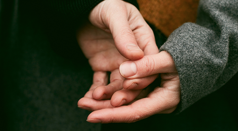 Two hands gently holding each other, one wearing a gray sleeve and the other a black sleeve.