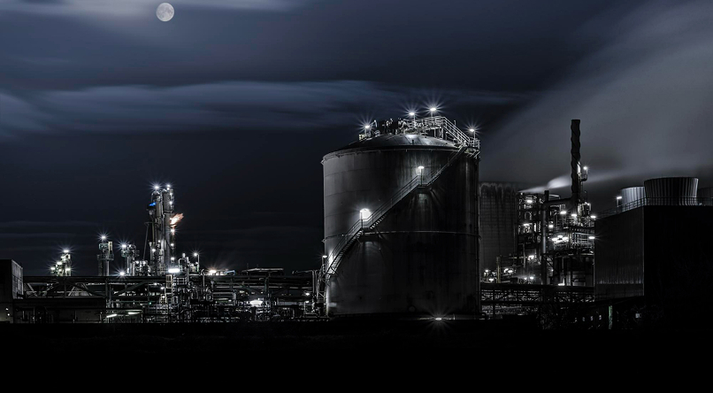 Industrial refinery complex illuminated at night under a cloudy sky with a visible full moon.