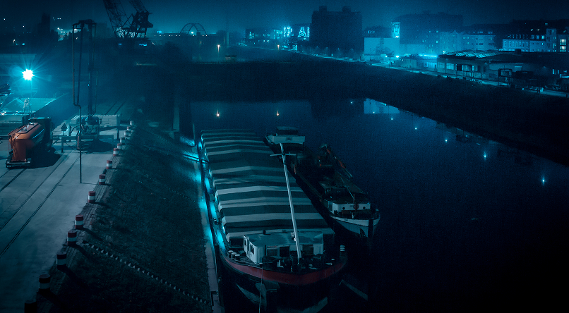 Large cargo ship docked at a port during nighttime with blue lighting and cranes in the background.