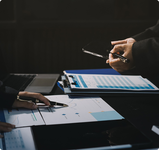 Two people discussing charts and graphs on printed documents with a laptop in the background.