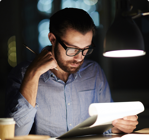 Man with glasses reviewing documents under a desk lamp in a dimly lit room.