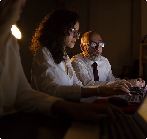 Two colleagues working closely on laptops in a dimly lit office, focused on the screen.