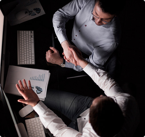 Two businesspeople shaking hands over a desk with charts and a keyboard, viewed from above.