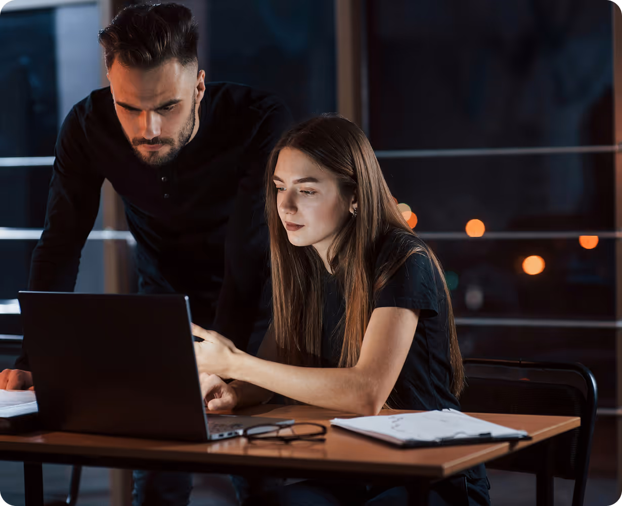 Man and woman working together at a laptop on a desk in a dimly lit office at night.