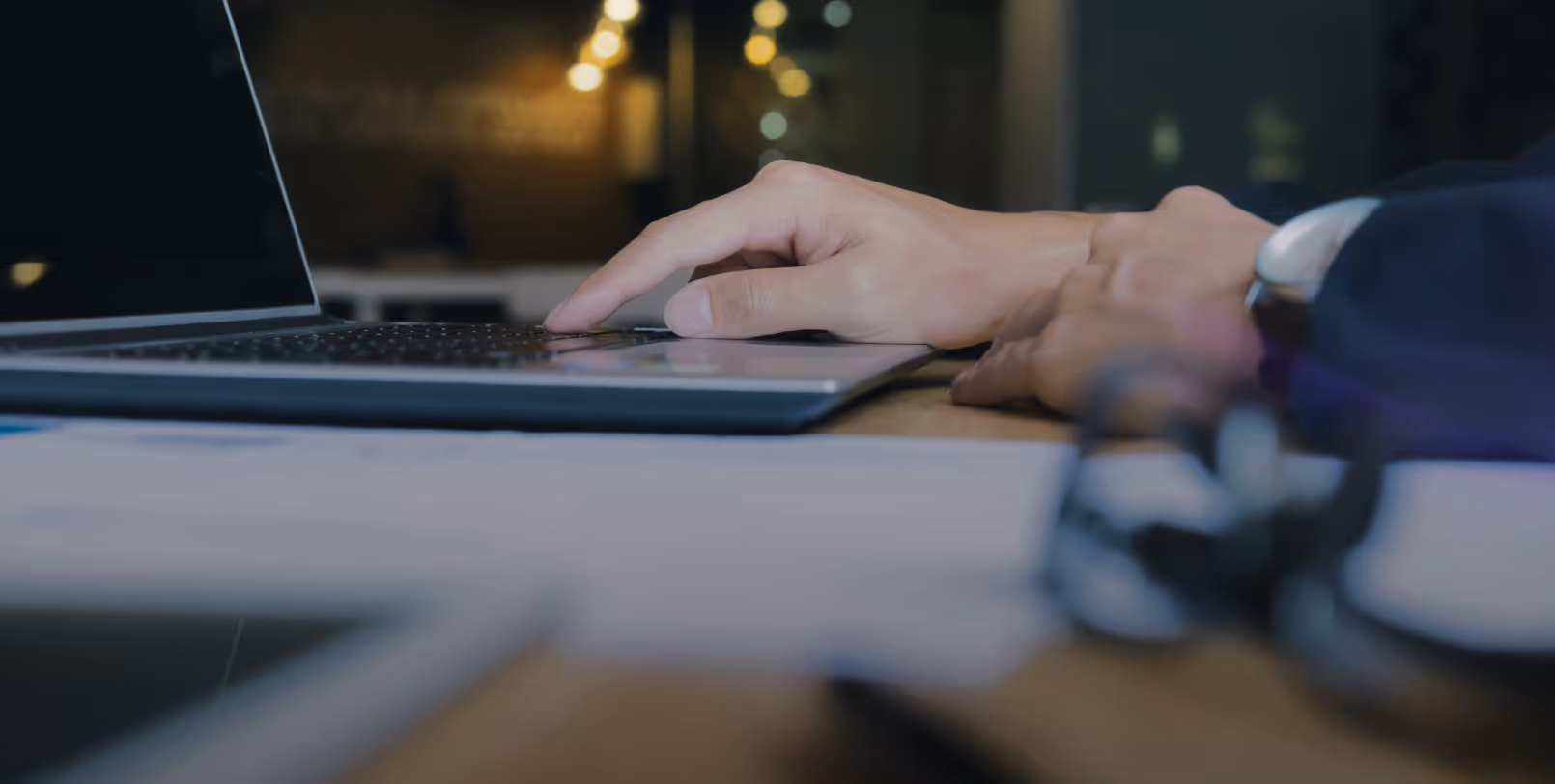 Close-up of a hand typing on a laptop keyboard on a wooden desk with blurred office background.