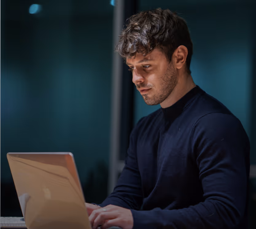 Young man focused on typing on a laptop in a dimly lit room.