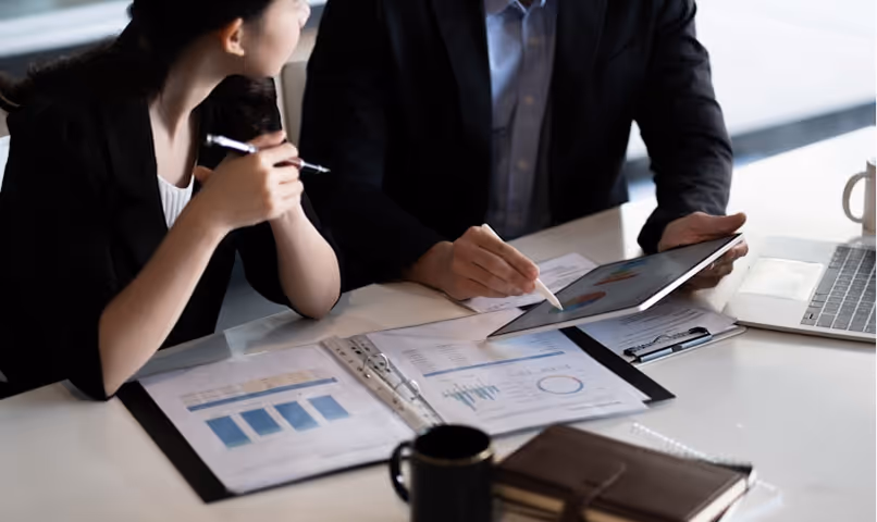 Two professionals discussing data charts on documents and a tablet at a desk with a laptop and coffee mug.