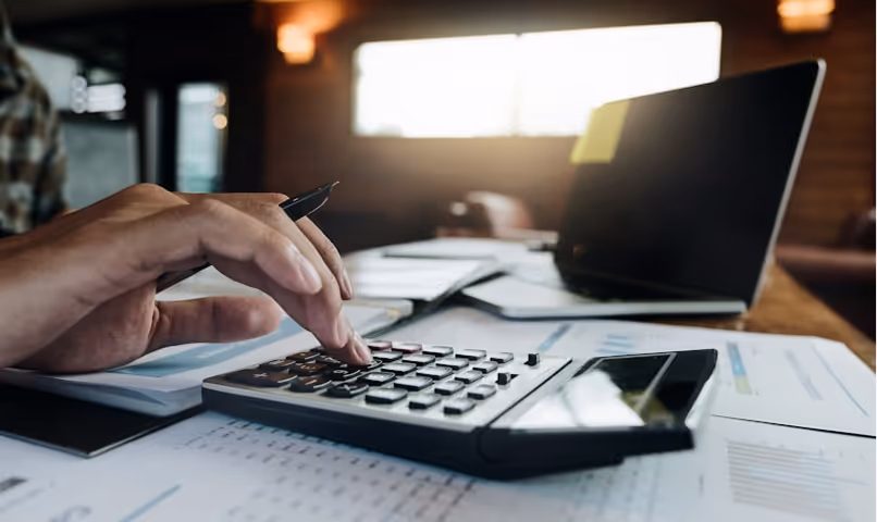 Person using a calculator with documents, a pen, and an open laptop on a wooden desk.