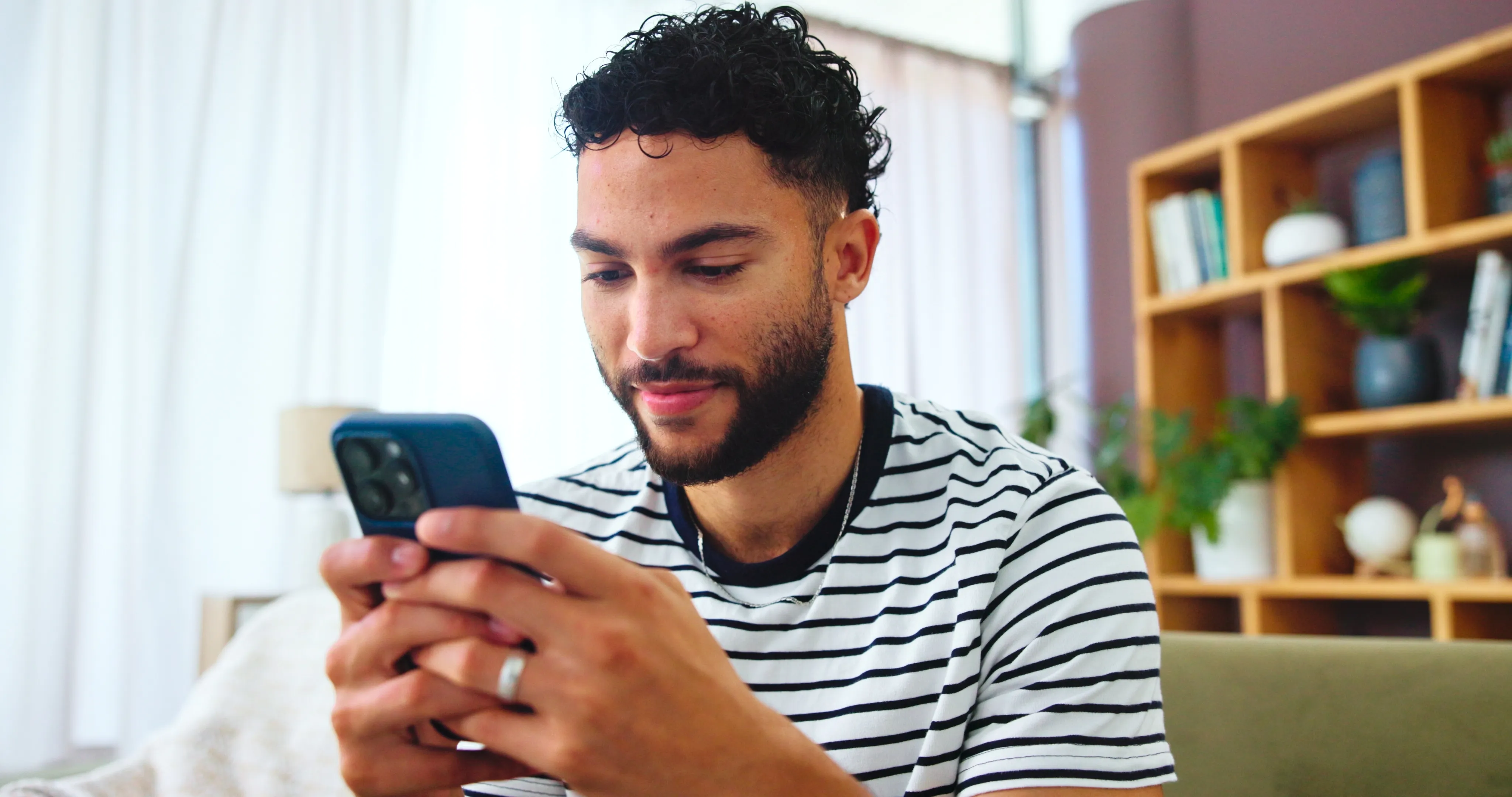 Mies katsoo ja käyttää puhelintaan kotona taustalla kirjahylly ja kasveja.