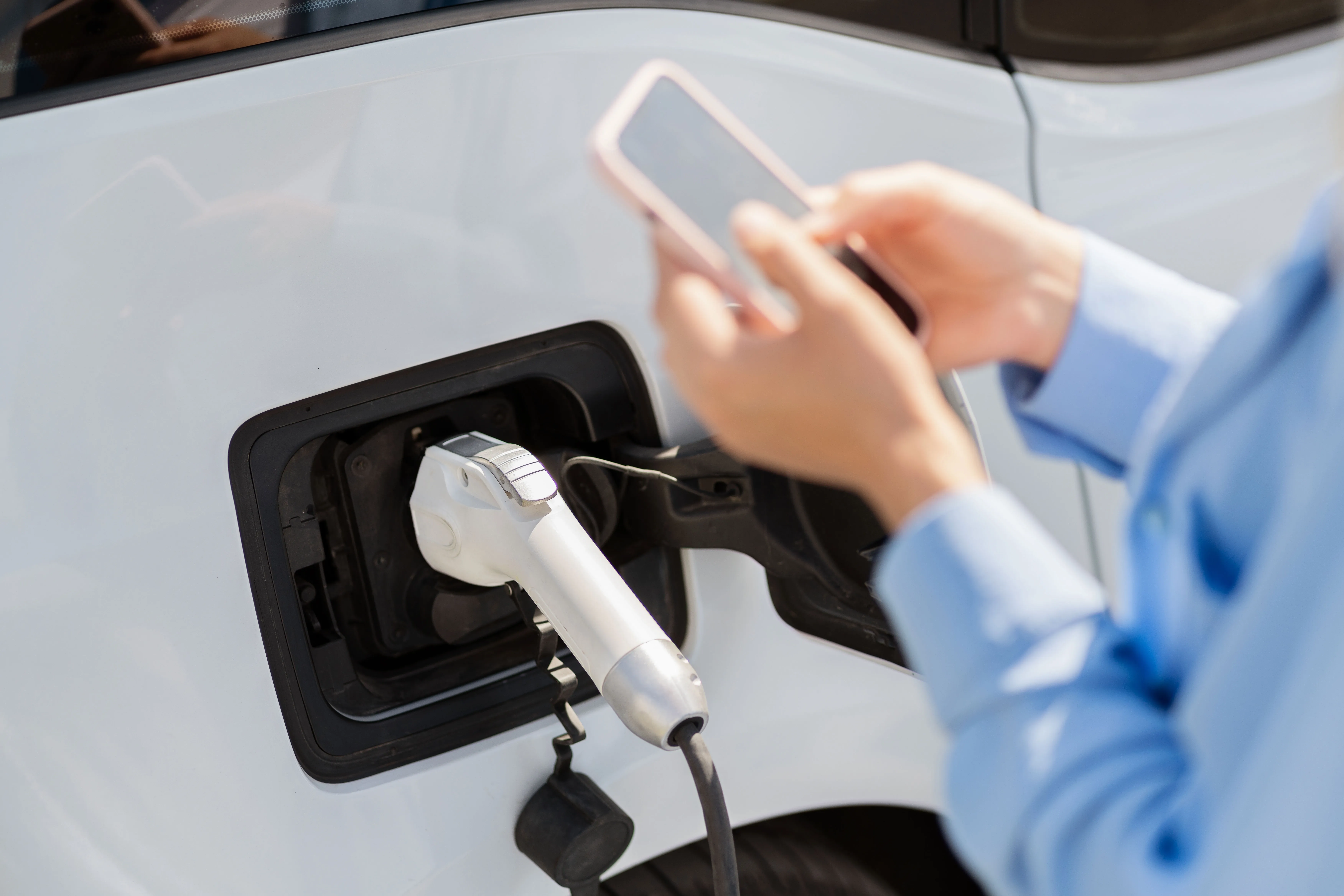 Henkilö pitelee älypuhelinta ja lataa sähköautoa valkoisella autolla.