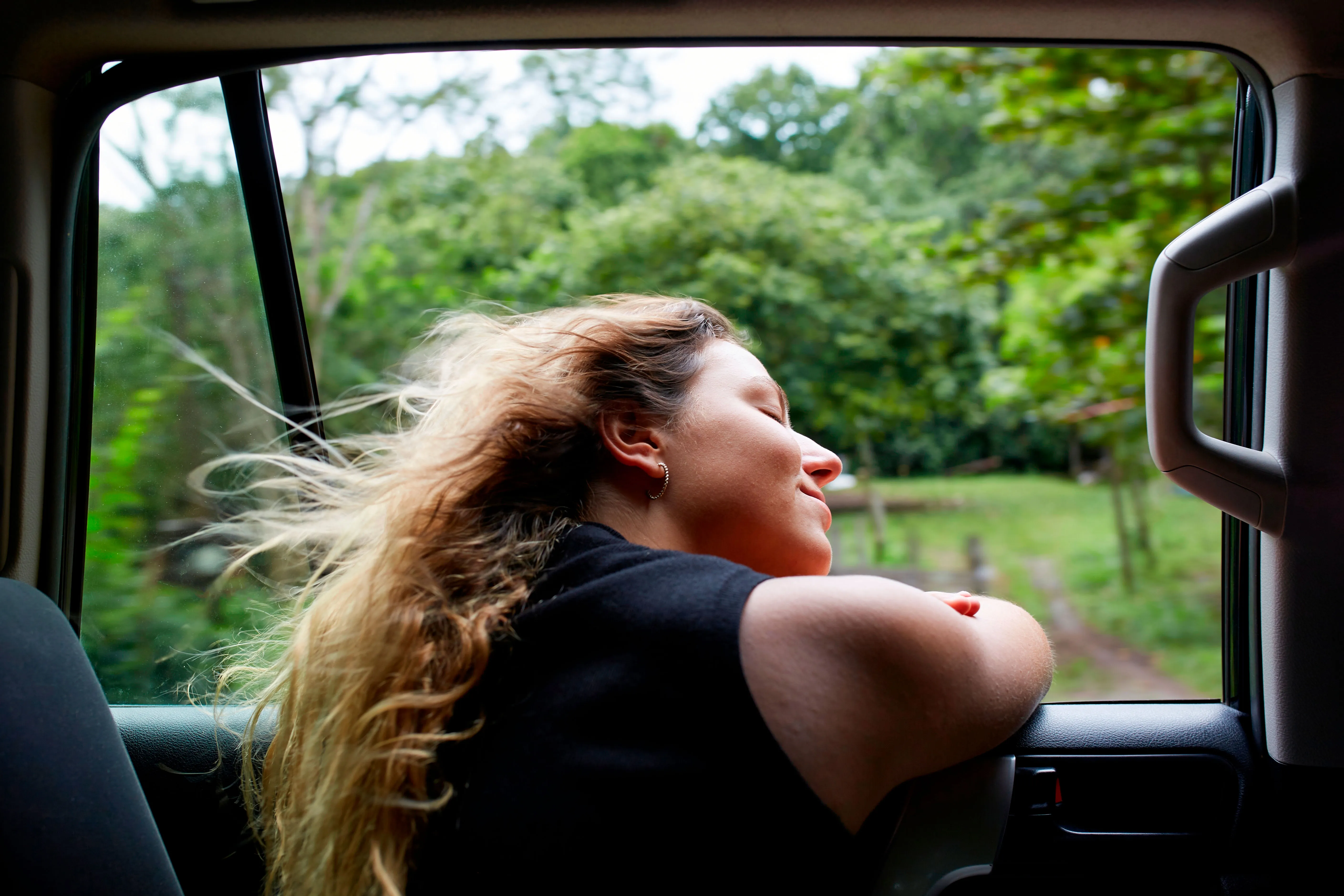 Nainen istuu autossa ikkunan vieressä ja nauttii ulkoilmasta suljetuin silmin hiukset tuulessa.