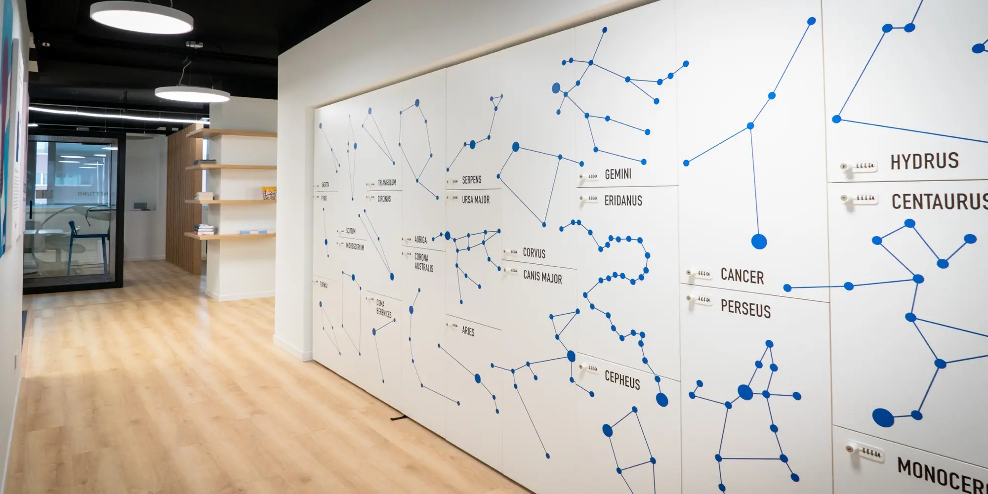 Corridoio di un ufficio con una grande parete bianca decorata con mappe di costellazioni astronomiche in blu, incluse Gemini, Eridanus, Cancer e Perseus