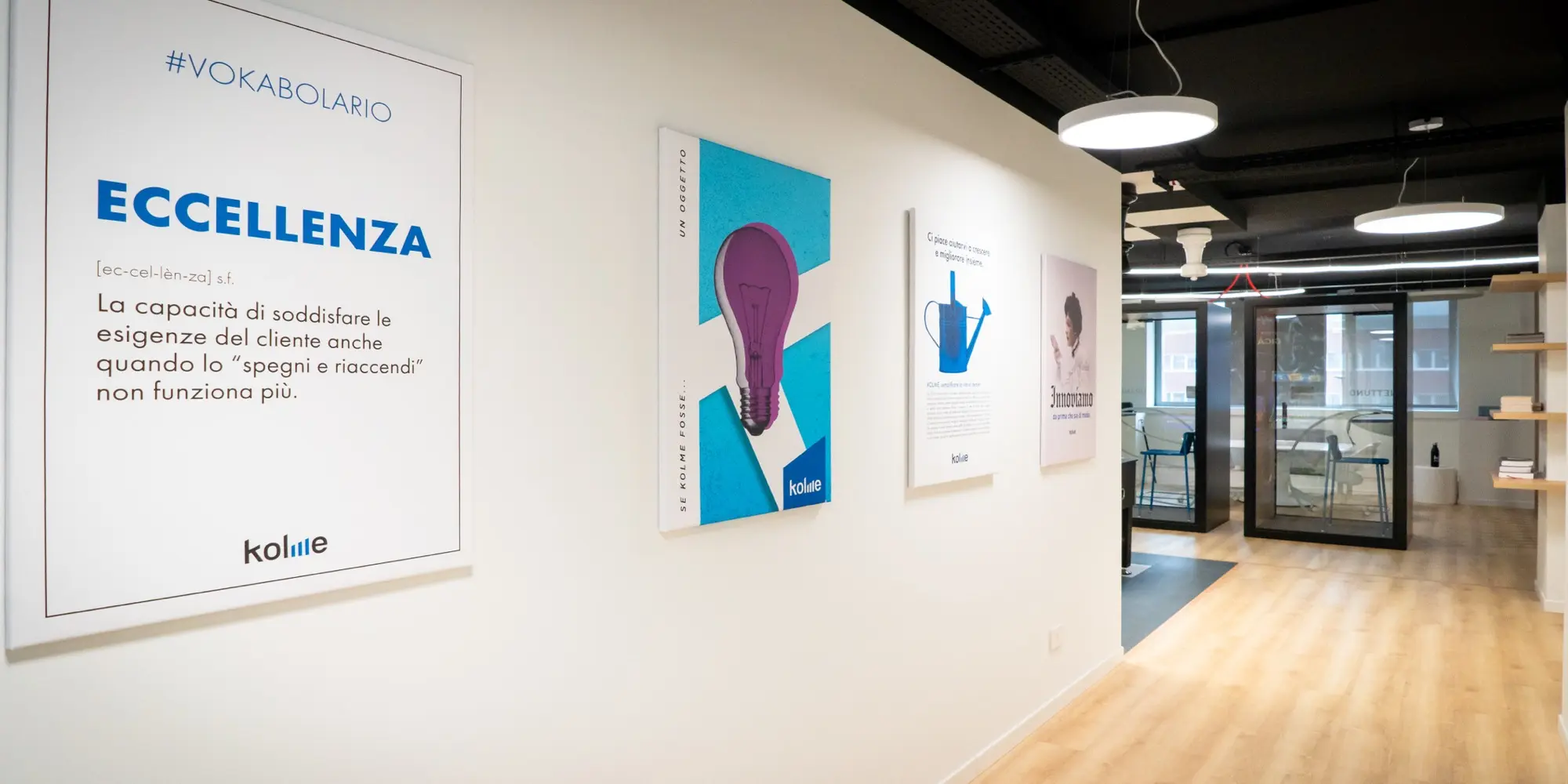 Corridoio con opere d’arte e poster incorniciati alle pareti, con parole come “ECCELLENZA” e vari design astratti. Il pavimento è in legno chiaro e sullo sfondo porte in vetro conducono a un’area riunioni.