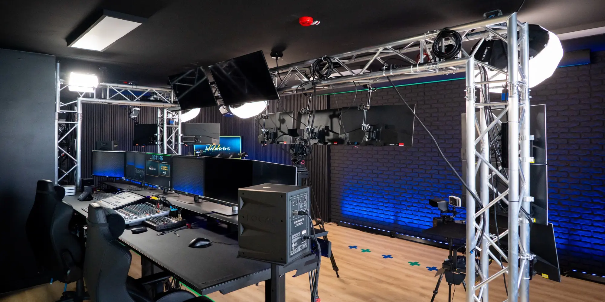 Studio di produzione video professionale con postazione di regia, schermi multipli, tralicci metallici per luci e telecamere, e parete di mattoni illuminata in blu