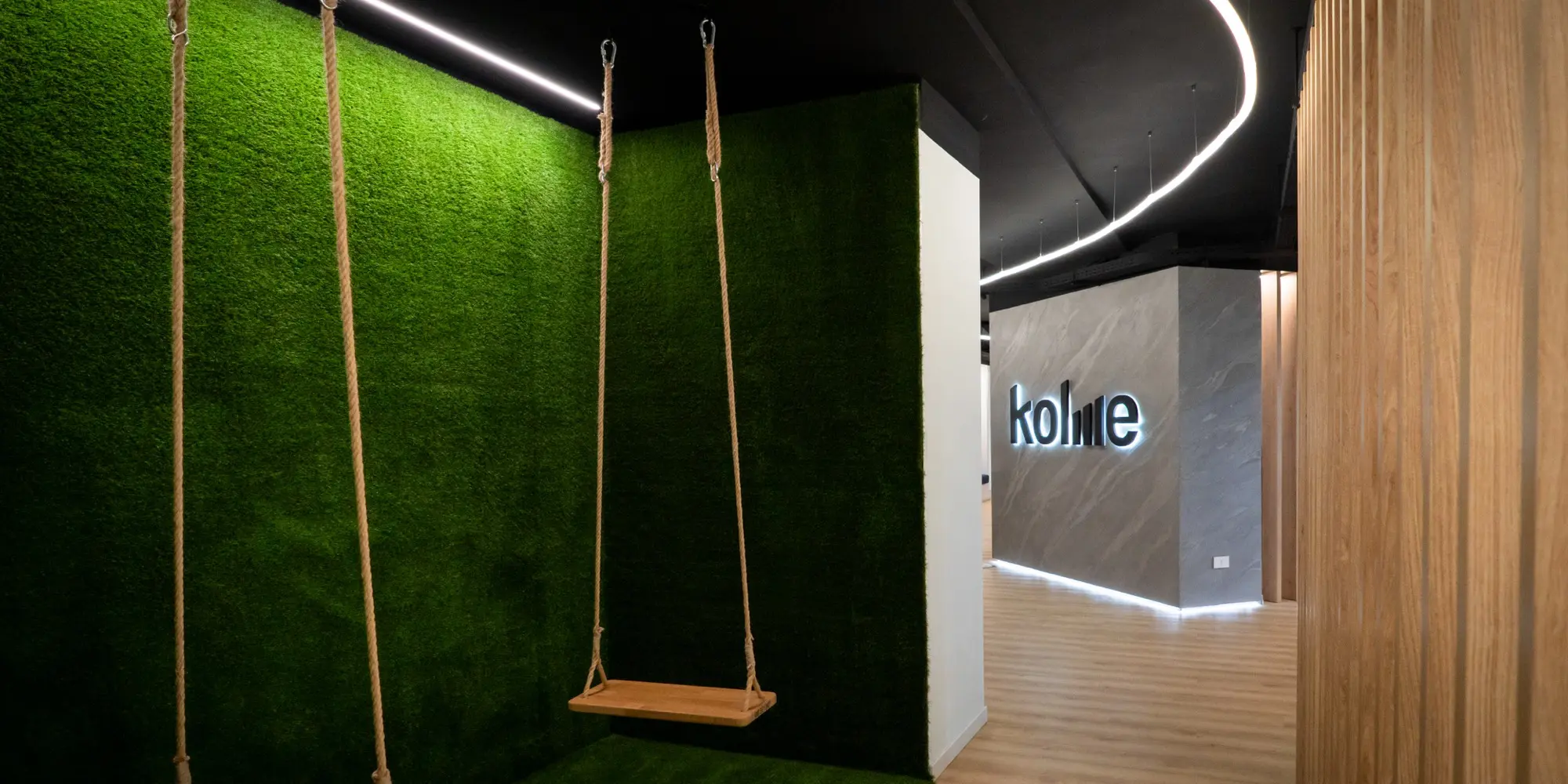 Ingresso degli uffici Kolme con altalena in legno appesa davanti a una parete di erba sintetica verde, e logo aziendale illuminato sullo sfondo