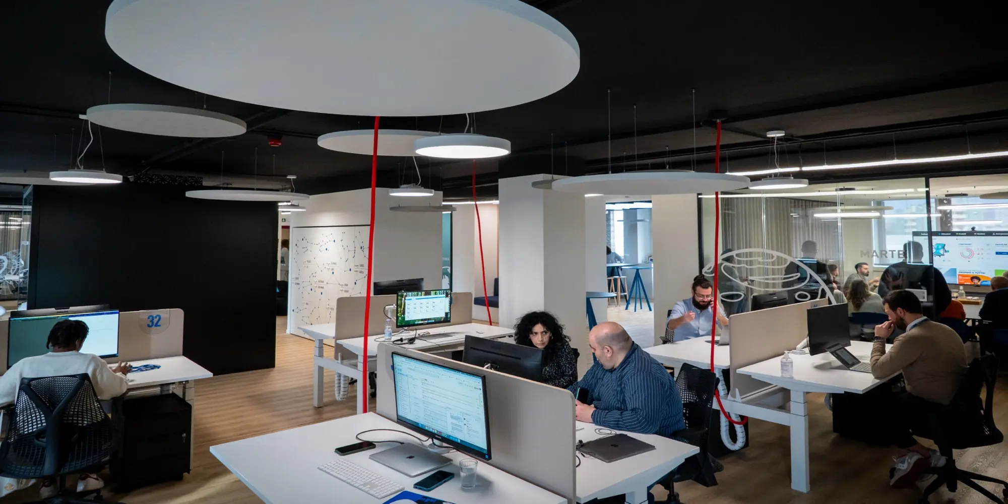 Open space di un ufficio con dipendenti al lavoro alle postazioni, pannelli acustici circolari a soffitto e colonne decorative rosse