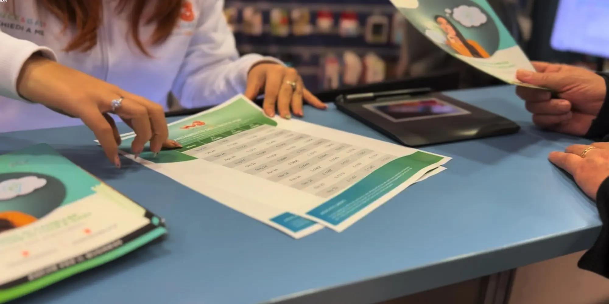 Operatrice di un punto vendita Windtre illustra materiale informativo a un cliente sul bancone, con brochure e schede tecniche colorate
