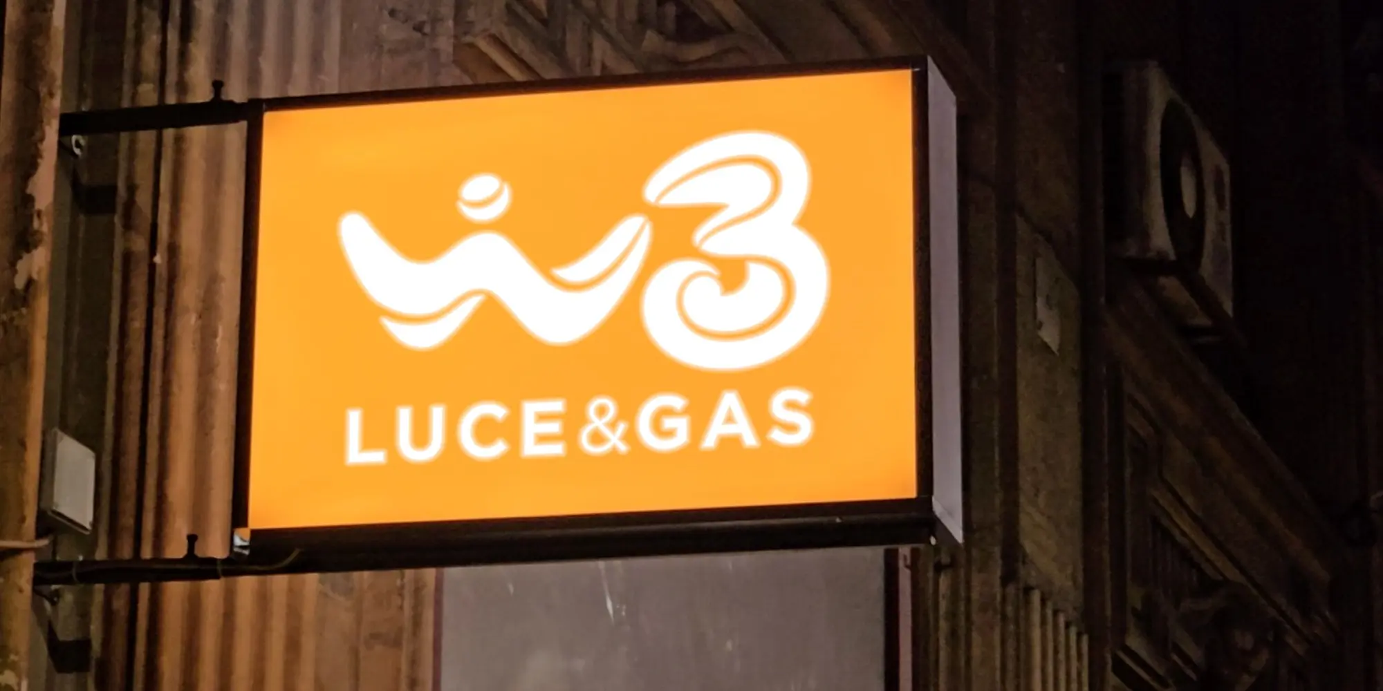 Insegna luminosa arancione Windtre Luce e Gas applicata alla facciata esterna di un edificio, fotografata di notte