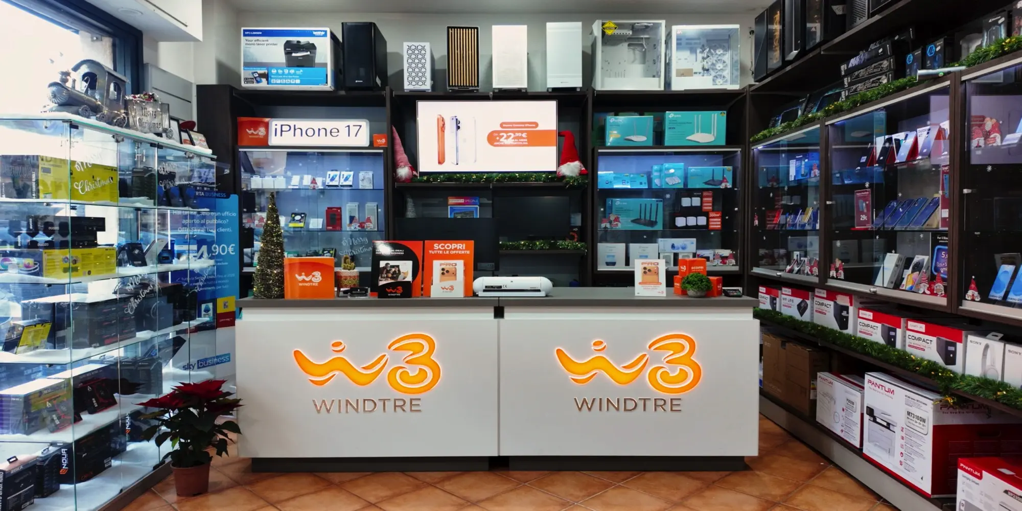 Interno di un punto vendita Windtre allestito per il Natale, con banco cassa brandizzato, espositori laterali di accessori e schermo promozionale