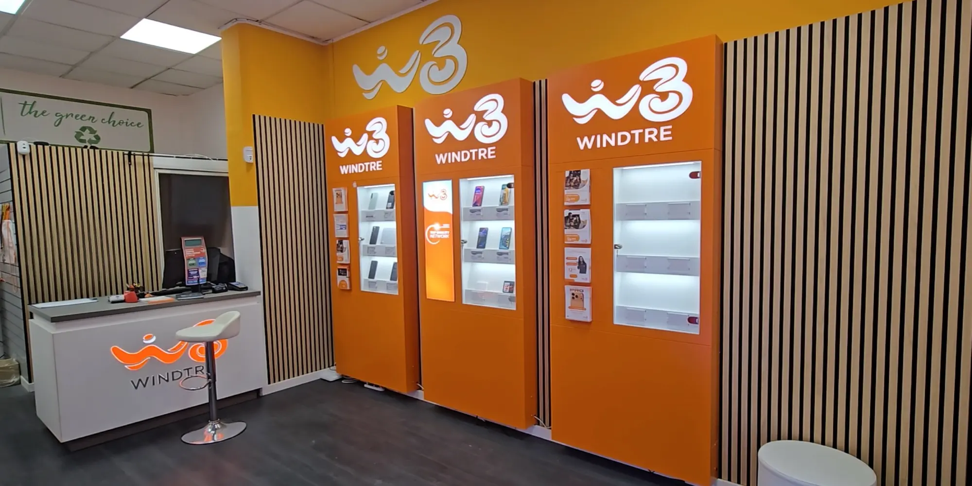 Interno di un negozio Windtre con pareti arancioni, colonne espositive illuminate con smartphone in mostra e bancone con logo del brand
