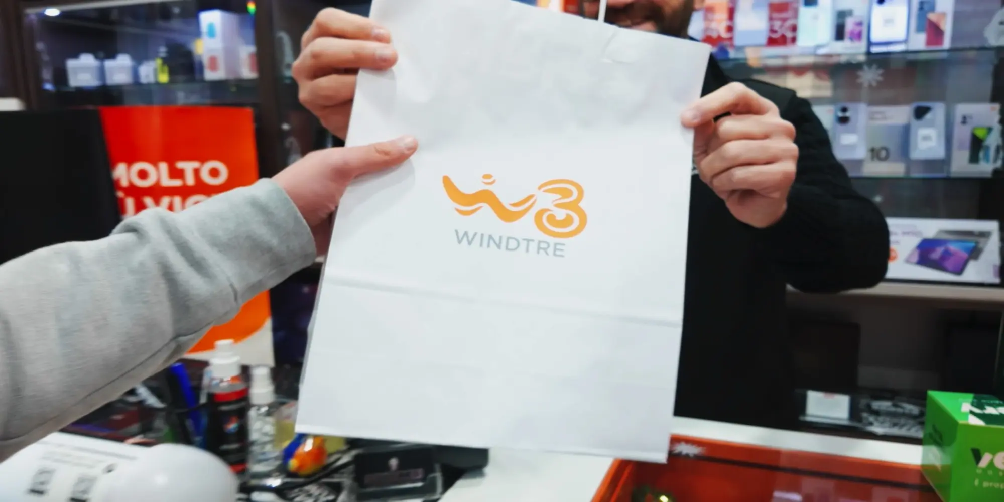 Operatore di un punto vendita Windtre consegna a un cliente una shopper bianca con il logo arancione del brand
