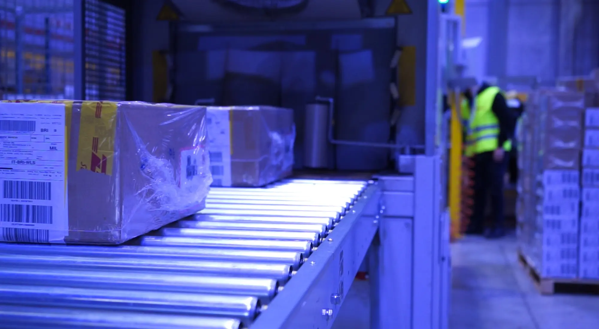 Pacco DHL avvolto in cellophane su un nastro trasportatore a rulli in un centro di smistamento, con operatore in gilet catarifrangente sullo sfondo