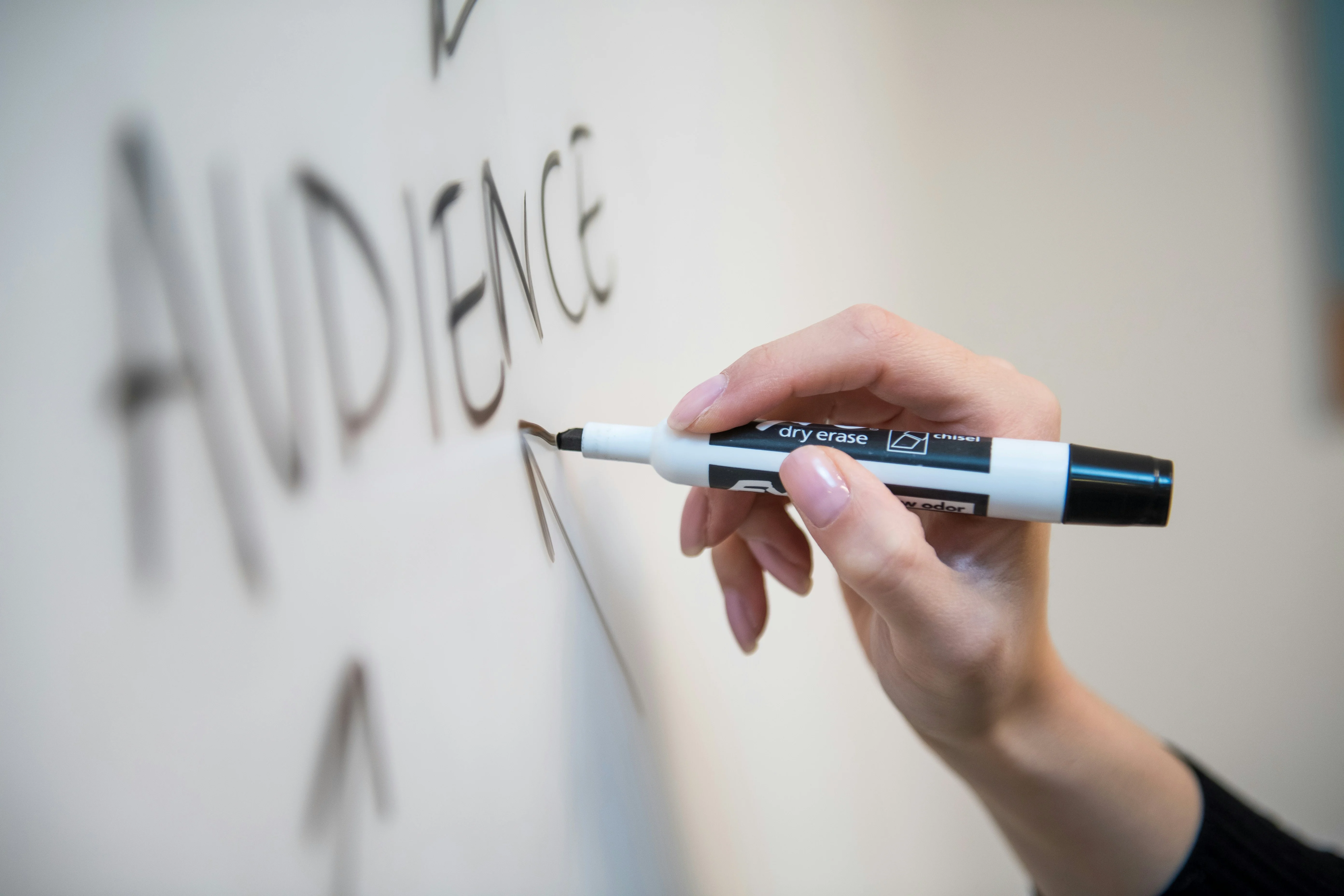 Hand holding a dry erase marker writing the word 'AUDIENCE' on a whiteboard with an arrow pointing up.